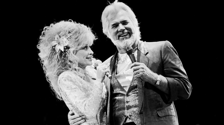 Kenny Rogers holds Dolly Parton by her waist as they both smile while singing.