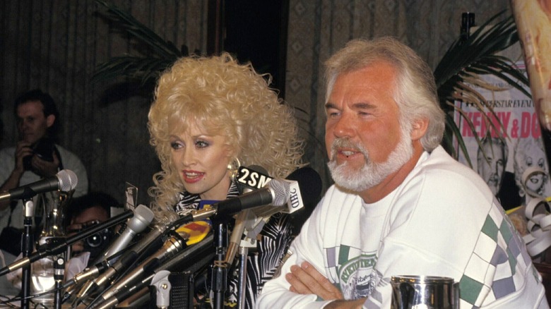 Dolly Parton and Kenny Rogers look away from the camera while sitting behind a sea of microphones.