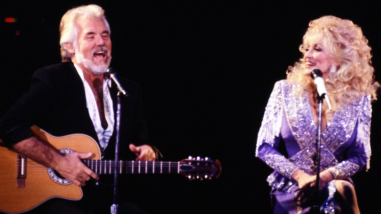 Kenny Rogers sings as a happy Dolly Parton looks at him while smiling.
