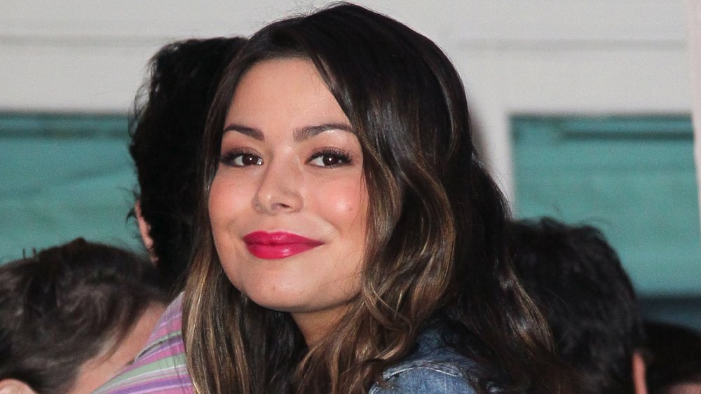Miranda Cosgrove in a jean jacket and pink lipstick, smiling while looking over her shoulder