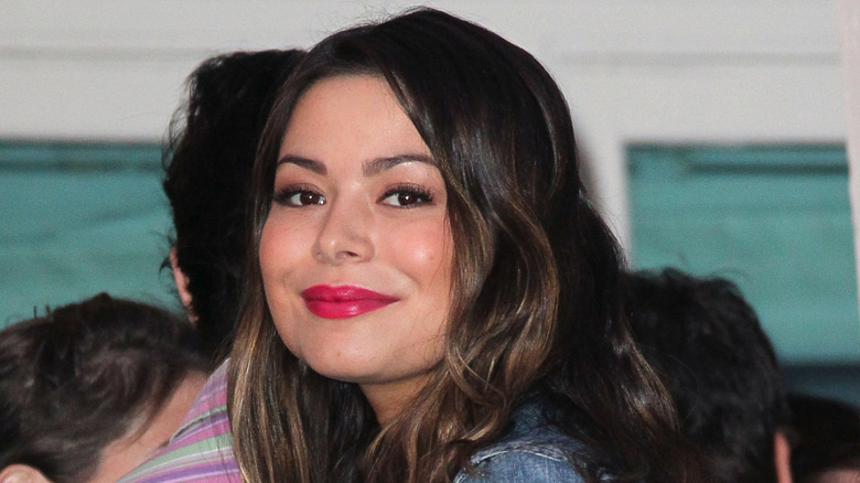 Miranda Cosgrove in a jean jacket and pink lipstick, smiling while looking over her shoulder