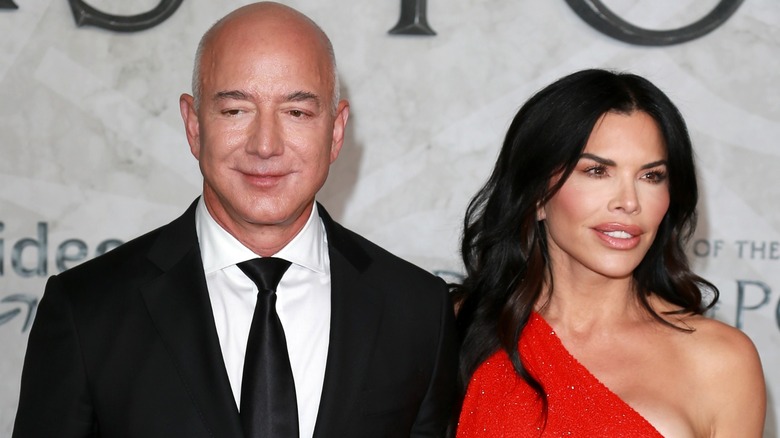 Sporting a one-shoulder red dress, Lauren Sanchez stands close to Jeff Bezos and looks away from the camera, as Jeff looks down.