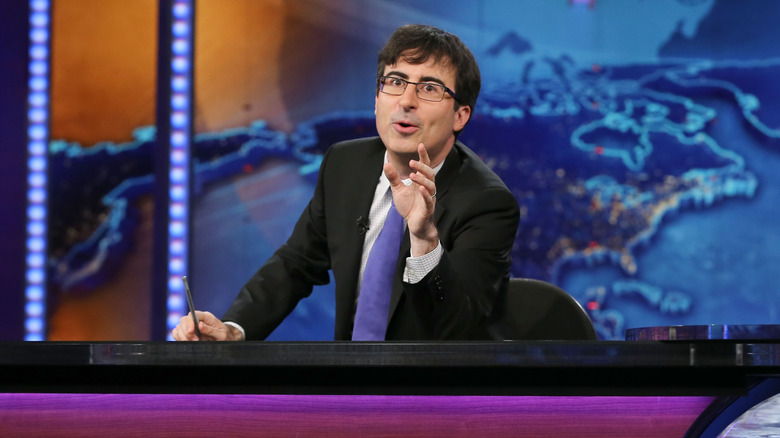 John Oliver speaking while sitting behind the desk on the set of The Daily Show