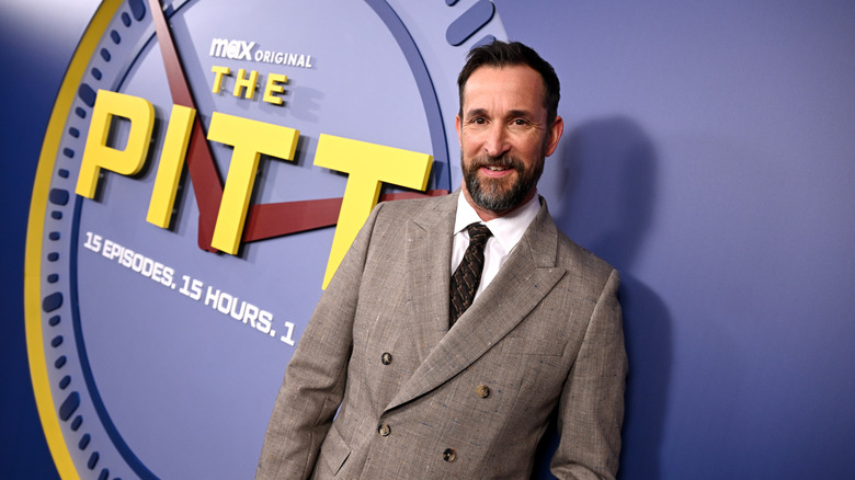 Noah Wyle wearing a tan suit standing in front of a backdrop for The Pitt