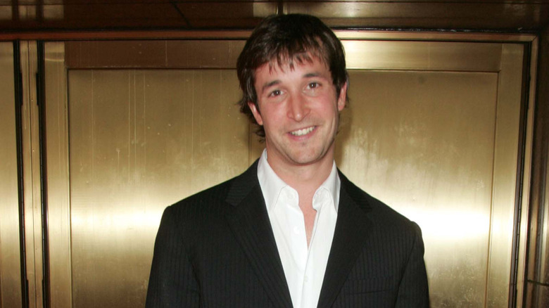Noah Wyle wearing a black pinstriped jacket and open collared white shirt