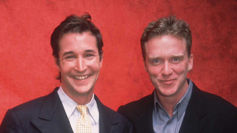 Noah Wyle wearung a black suit jacket and yellow patterned tie standing with Anthony Michael Hall