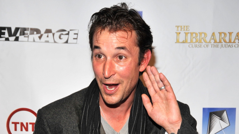 Noah Wyle holding a hand to his ear while wearing a grey jacket and black striped scarf