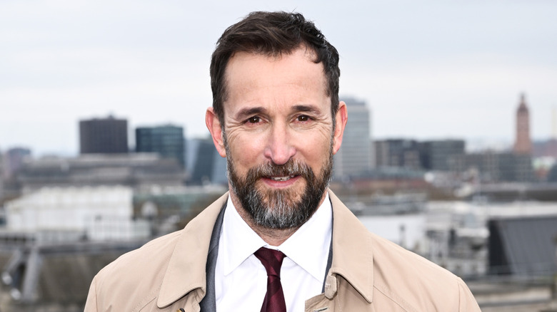 Noah Wyle wearing a tan trenchcoat, grey suit jacket, white collared shirt and maroon tie