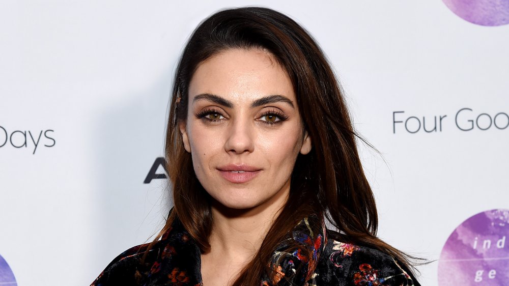 Mila Kunis in a black printed top, with a small smile, posing at an event