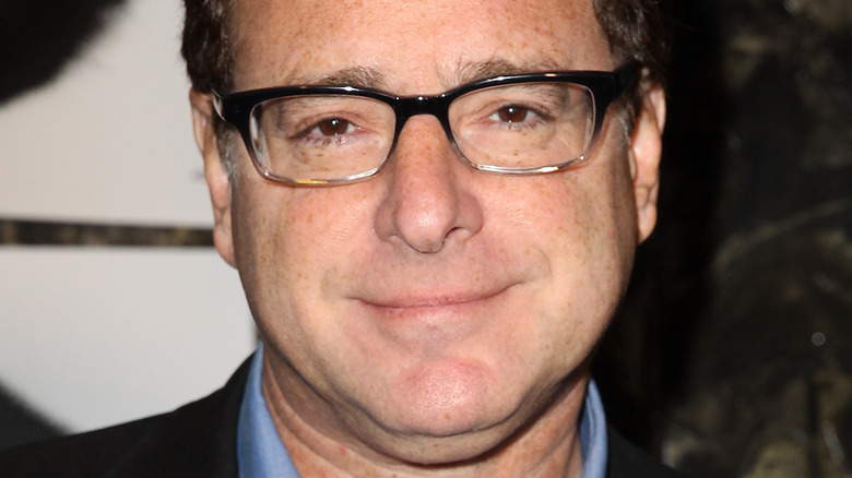 Bob Saget wearing black-framed glasses, smiles at camera