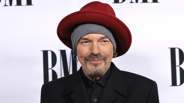 Billy Bob Thornton wearing a black suit jacket, black collared shirt, grey scarf around his head, and a red wide-brimmed hat
