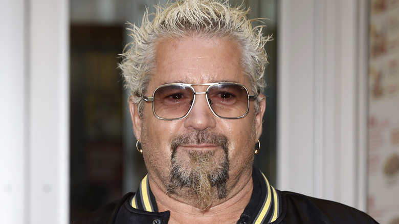 Guy Fieri posing for cameras in a black varsity jacket and tinted glasses