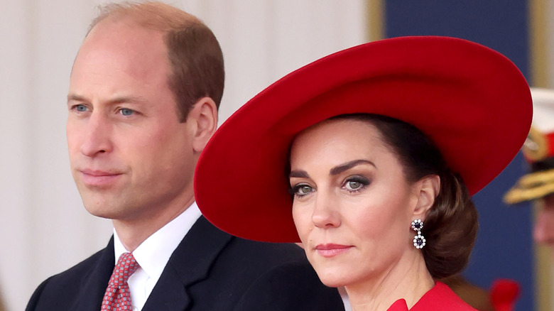 Prince William standing next to Kate Middelton who is wearing a large red hat and dress