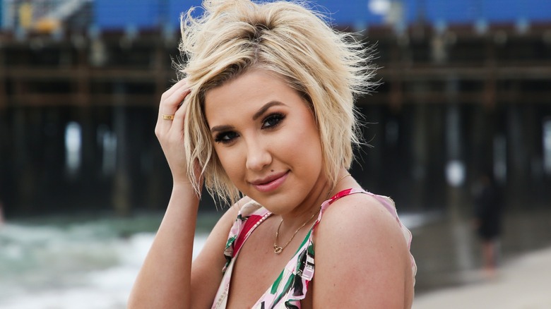 Sporting a short haircut, Savannah Chrisley tilts her head to the side as she brushes her hair with one hand.