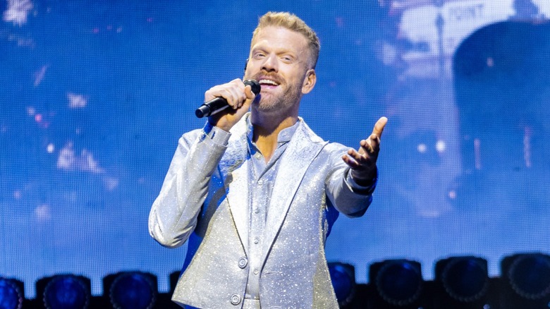 Wearing a shimmery suit, Scott Hoying sings while extending an open palm.