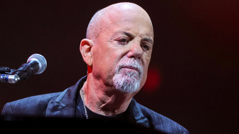 Billy Joel furrowing eyebrows and looking downward beside a microphone