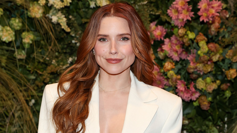 Sophia Bush smiling in a white jacket against foliage