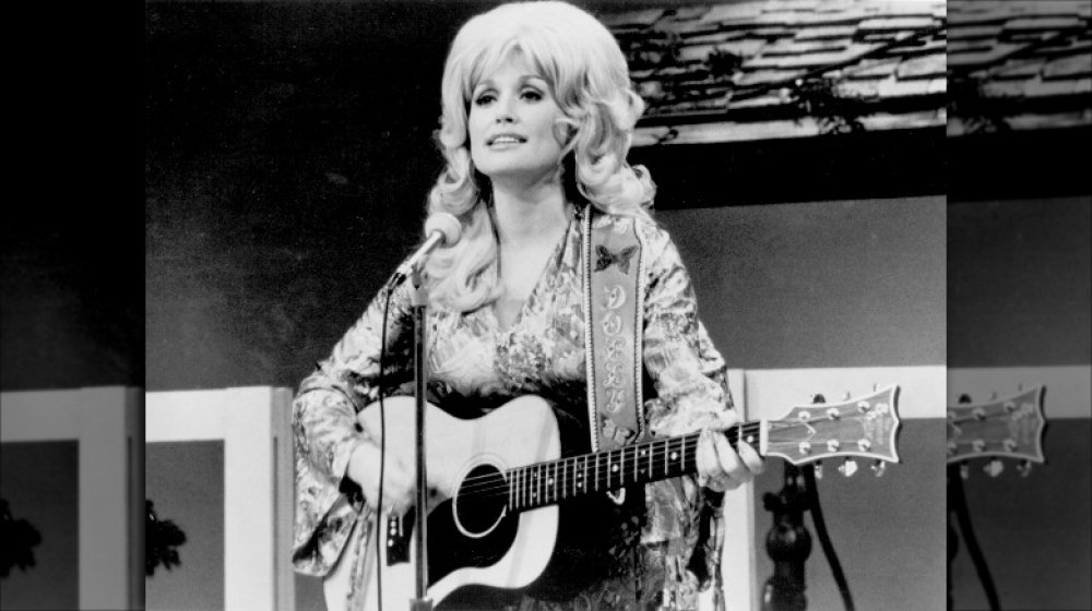 Black-and-white photo of Dolly Parton singing and playing guitar in 1974 at age 28