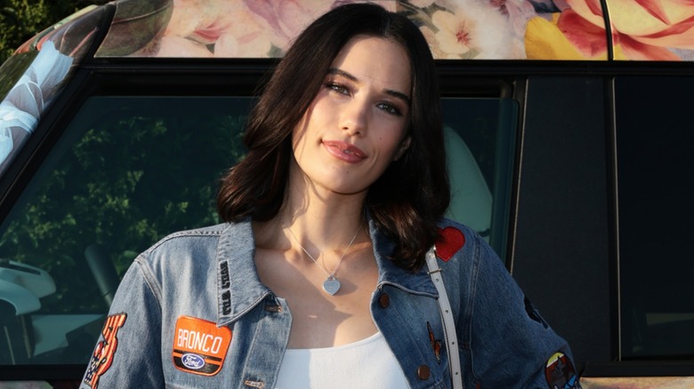 Wearing a denim and tank top combo, Ella Travolta smiles gently with her head tilted to the side.