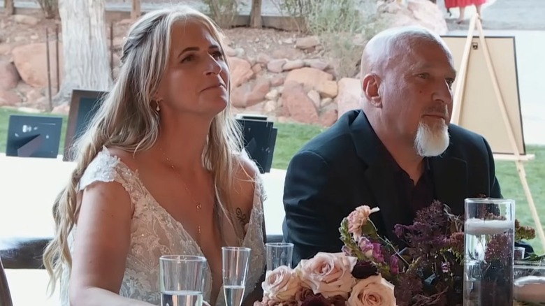Christine Brown and David Woolley look away from the camera while seated at a table with wedding decor