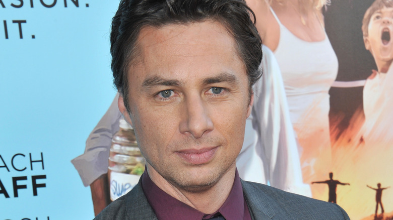 Zach Braff, smirking, promoting "Wish I Was Here," standing in front of the movie poster in 2014