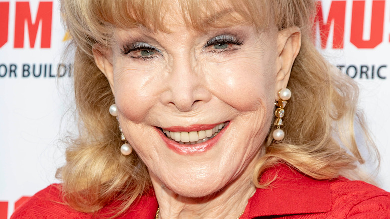 Barbara Eden smiles in a red outfit and pearl earrings