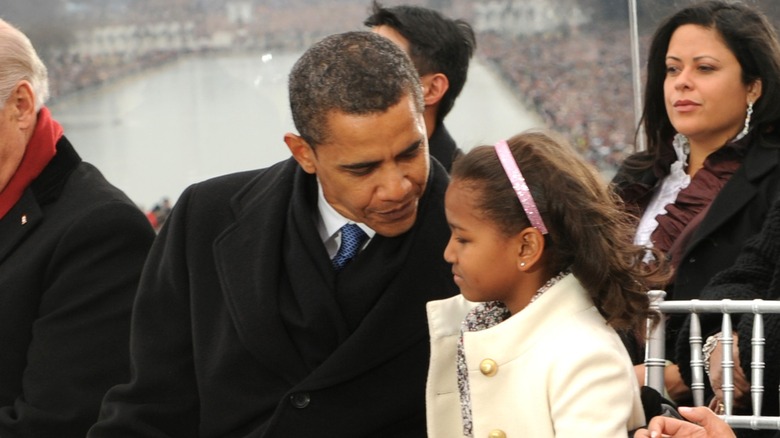 Barack Obama leans and talks to young Sasha Obama, who's wearing a cream coat and a pink headband.