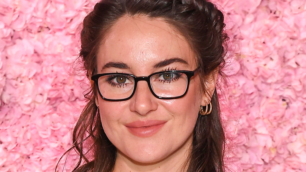 Shailene Woodley standing against a wall of pink roses wearing glasses