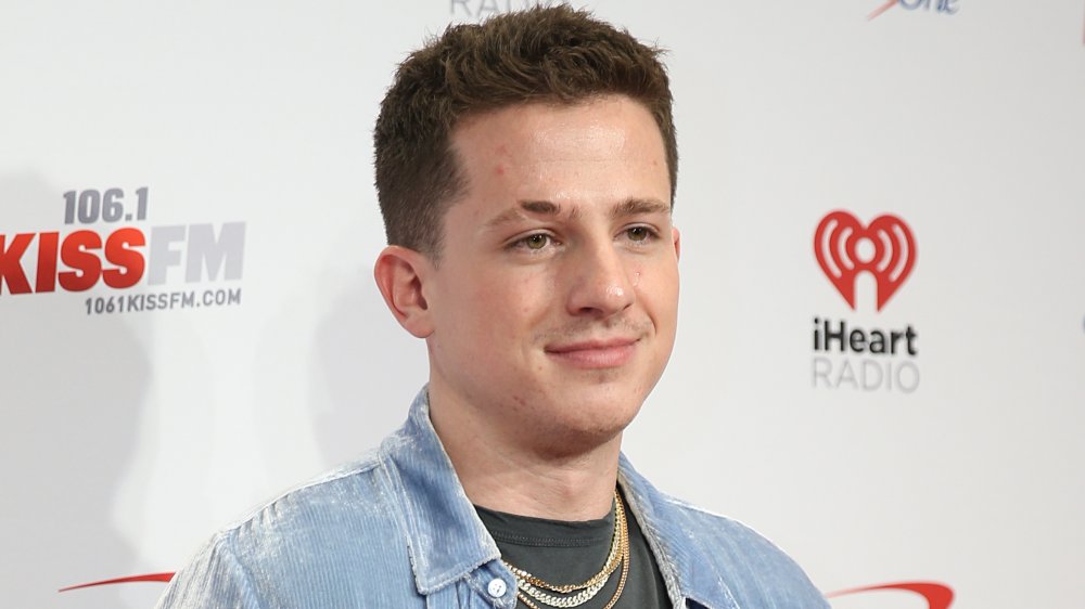 Charlie Puth in a light blue corduroy button down, black t-shirt, and chains, posing with a neutral expression at an iHeartRadio event