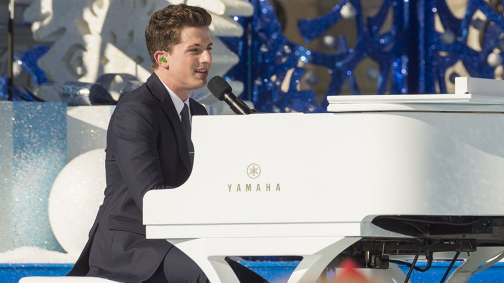 Charlie Puth in a navy suit, singing and playing a white piano