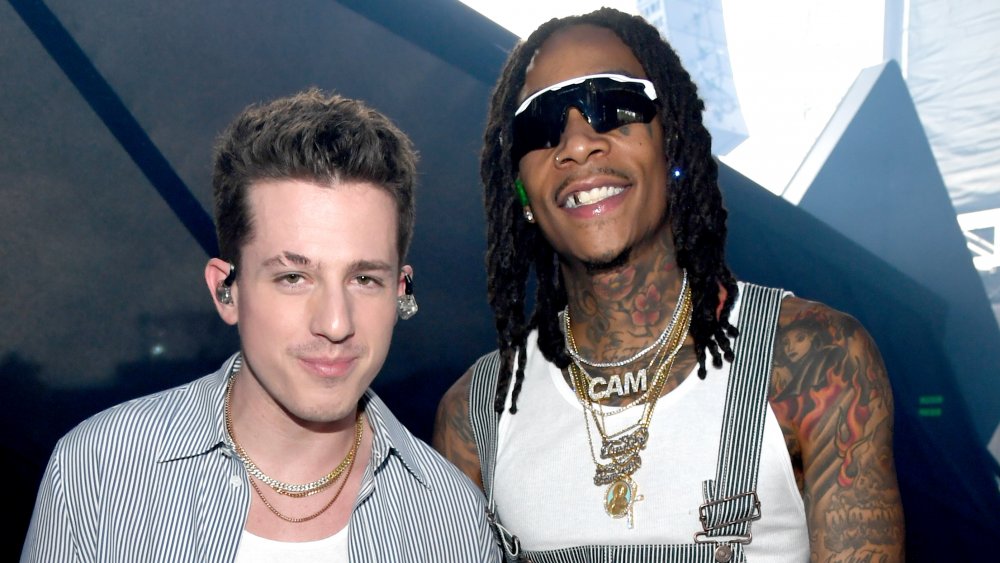 Charlie Puth in a striped button down with a white t-shirt, and Wiz Khalifa in a white tank top, sunglasses, and striped overalls, smiling and posing together backstage