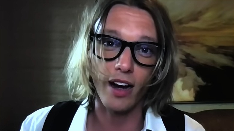 Jamie Campbell Bower wearing thick glasses, speaking