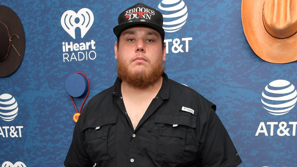 Luke Combs in a black button up and cap, posing with a serious expression at an iHeartRadio event