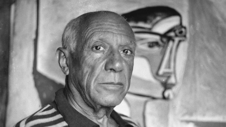 Pablo Picasso looks at camera with painting in background