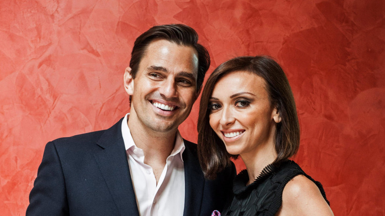 Bill Rancic and Giuliana Rancic smile as they pose against a red backdrop.
