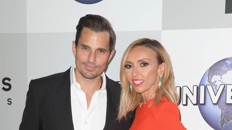 Bill Rancic gives the camera a piercing stare while Giuliana Rancic, dressed in a fierce orange outfit, smiles.
