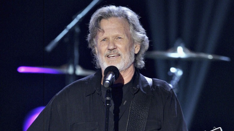 Kris Kristofferson, wearlng an all-black look, sings into a microphone.