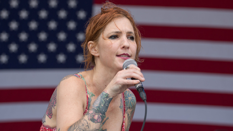 Casey Kristofferson, wearing smokey eye makeup, sings into a microphone in front of an American flag backdrop.