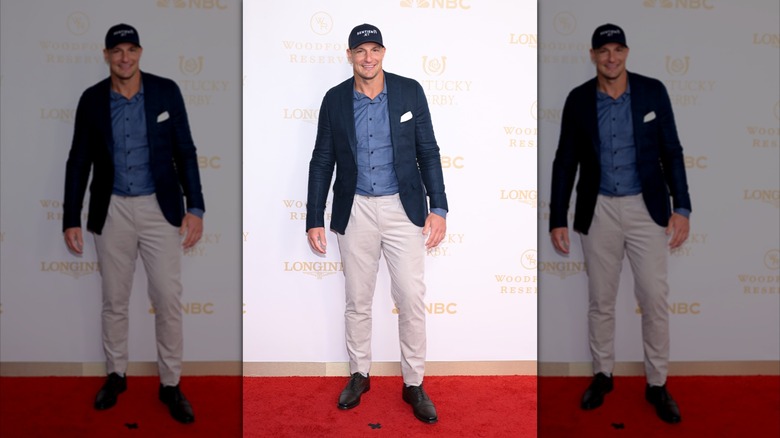 Rob Gronkowski smiling while wearing a dark baseball cap, matching sport coat, and khakis on the red carpet at Kentucky Derby.
