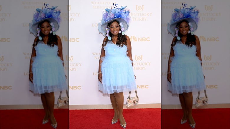Star Jones smiling and posing at the Kentucky Derby in a blue ruffled dress and hat covered with blue and purple flowers.