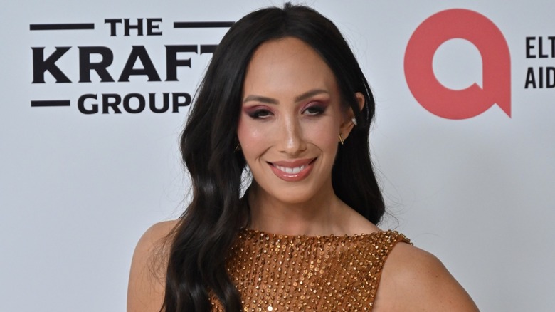 Cheryl Burke, wearing soft makeup and a sequined outfit, smiles while gazing downward.