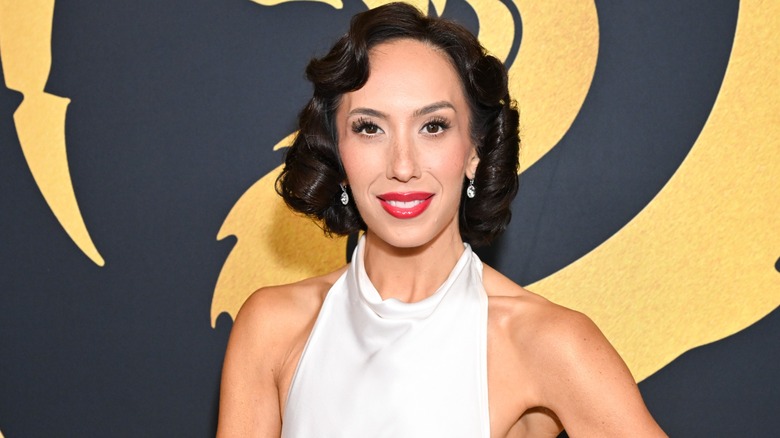 Cheryl Burke smiles radiantly while wearing short, curly hair and a white outfit.