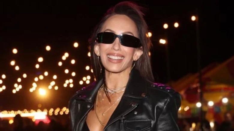 Cheryl Burke smiles while posing against a background of night lights.