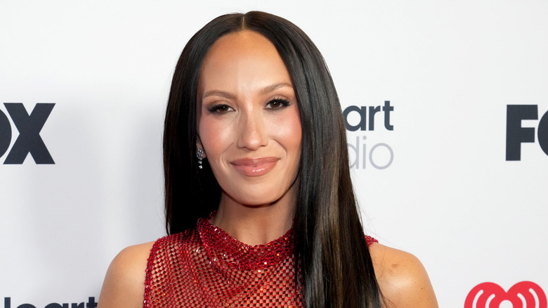 Cheryl Burke smiles softly while wearing her hair parted and a see-through red outfit.