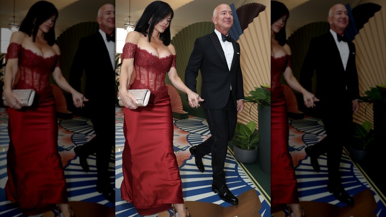 Wearing a red wine gown with lacy and opaque detail, Lauren Sánchez walks while holding hands with Jeff Bezos.