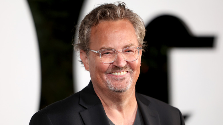 Matthew Perry smiling while wearing glasses, a black jacket, and black t-shirt