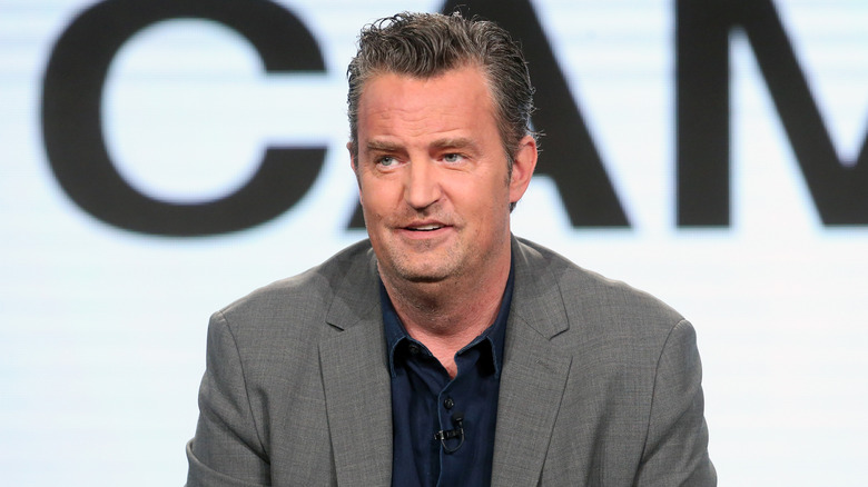 Matthew Perry speaking at an event wearing a grey jacket and blue button down shirt