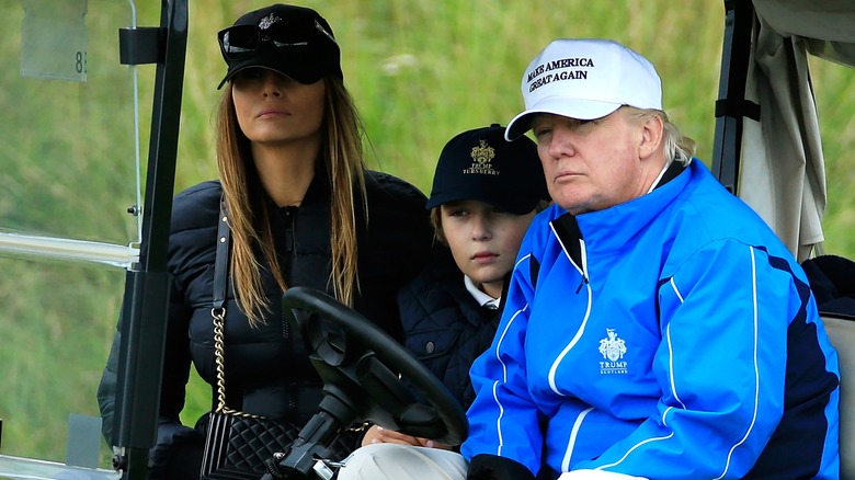 Young Barron Trump sits between Melania and Donald Trump in a golf cart.