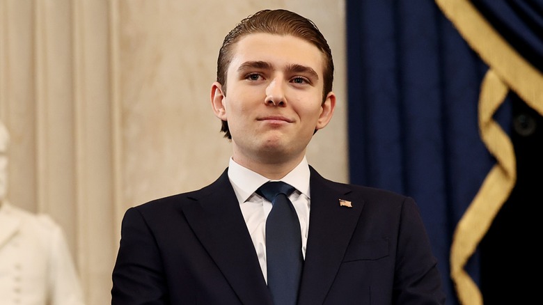 Dressed in a black suit with an American flag pin, Barron Trump smirks as he looks away from the camera.
