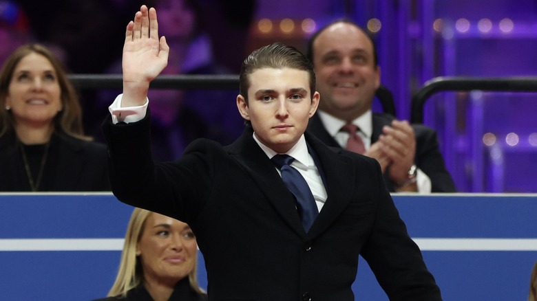 Sporting a black overcoat, a white shirt, and a navy blue tie, Barron Trump waves to a crowd.